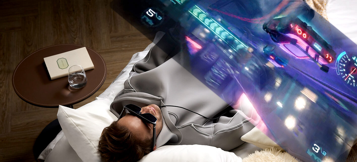 一名男子躺在床上，配戴 ROG XREAL R1 電競 AR 眼鏡與 ROG Ally 掌機享受沉浸式遊戲體驗